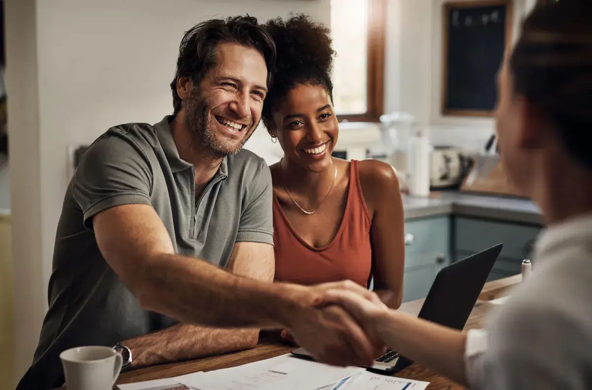 A couple sitting at a table, one of them shaking hands with an advisor A couple sitting at a table, one of them shaking hands with an advisor