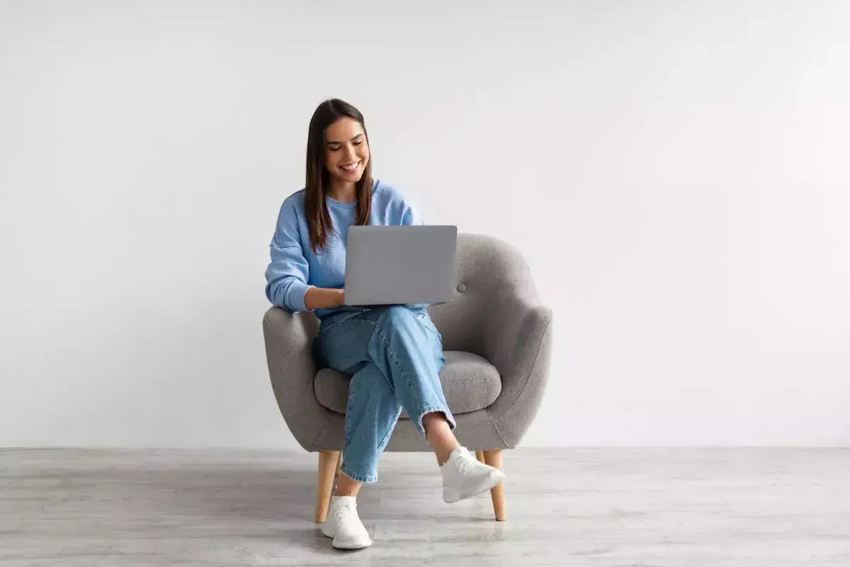 Young woman in blue outfit sitting in gray armchair, smiling while using a laptop.